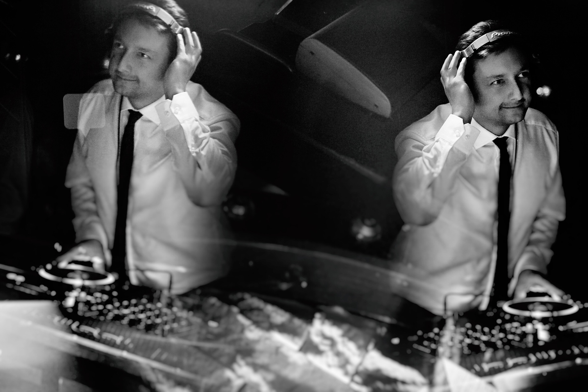 Czarno-białe zdjęcie DJ-a w białej koszuli i krawacie, z słuchawkami na głowie, miksującego muzykę na profesjonalnym sprzęcie DJ-skim, z efektem podwójnej ekspozycji.