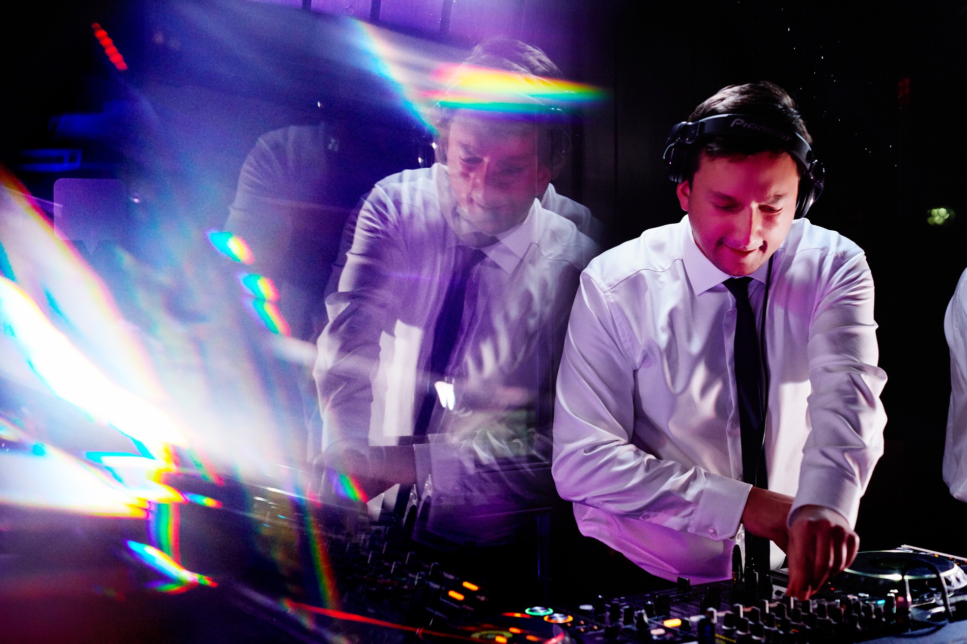 DJ w białej koszuli i słuchawkach Pioneer miksuje muzykę na konsolecie, z widocznym efektem rozszczepienia światła.