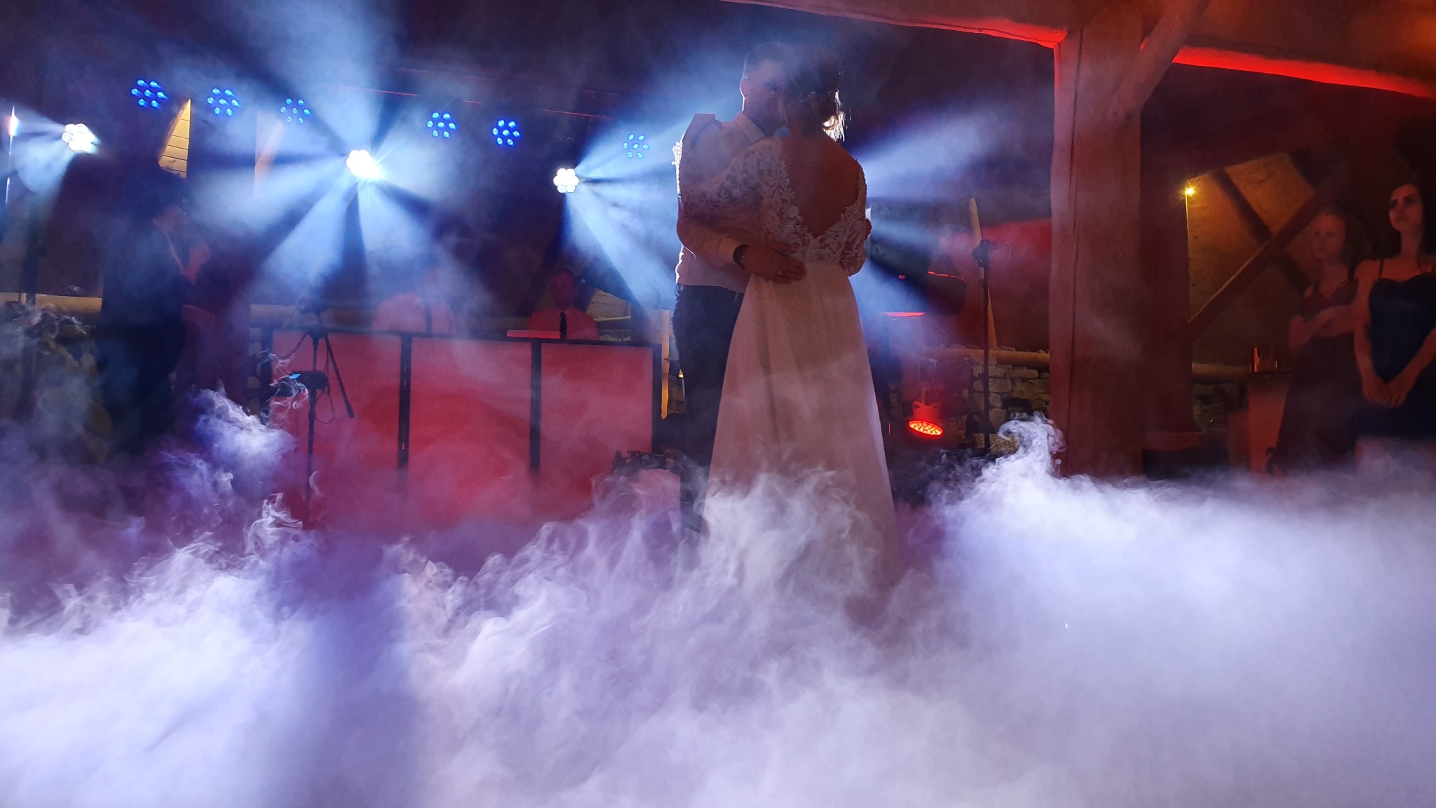 Para młoda tańczy w dymie na weselu, oświetlenie sceniczne w tle, DJ za konsoletą, drewniana konstrukcja sufitu.