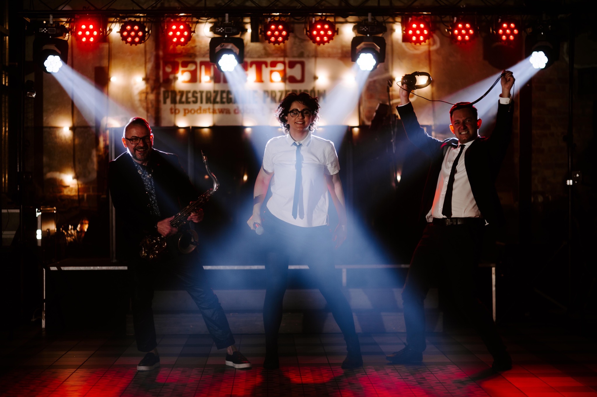 Zespół muzyczny w trakcie występu na scenie: saksofonista, wokalistka z mikrofonem i mężczyzna z słuchawkami, oświetlenie sceniczne i czerwone światła.