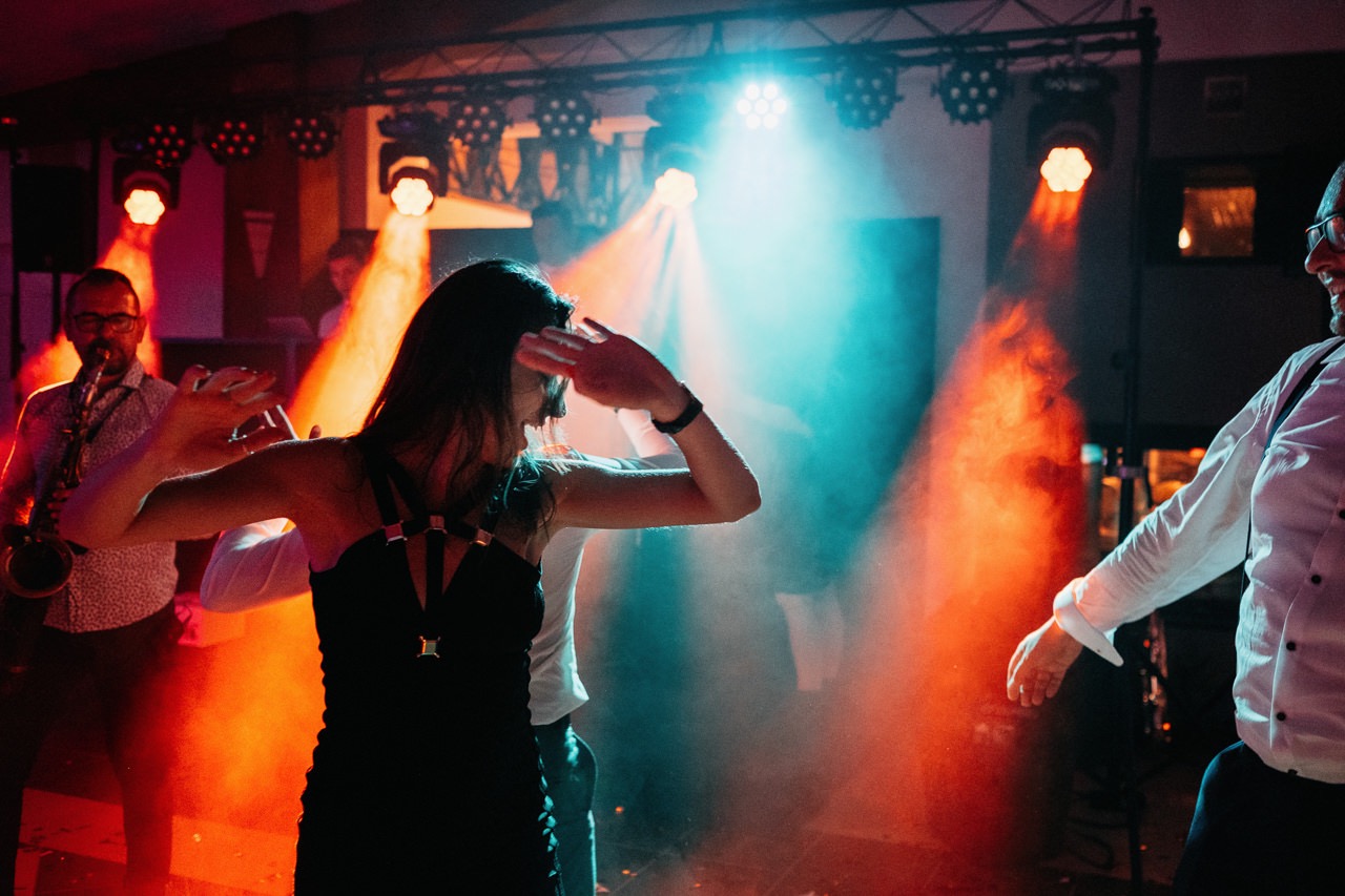 Parkiet taneczny z oświetleniem scenicznym: kobieta w czarnej sukience tańczy, w tle mężczyzna gra na saksofonie, dynamiczne światła w kolorach pomarańczowym i niebieskim tworzą atmosferę imprezy.