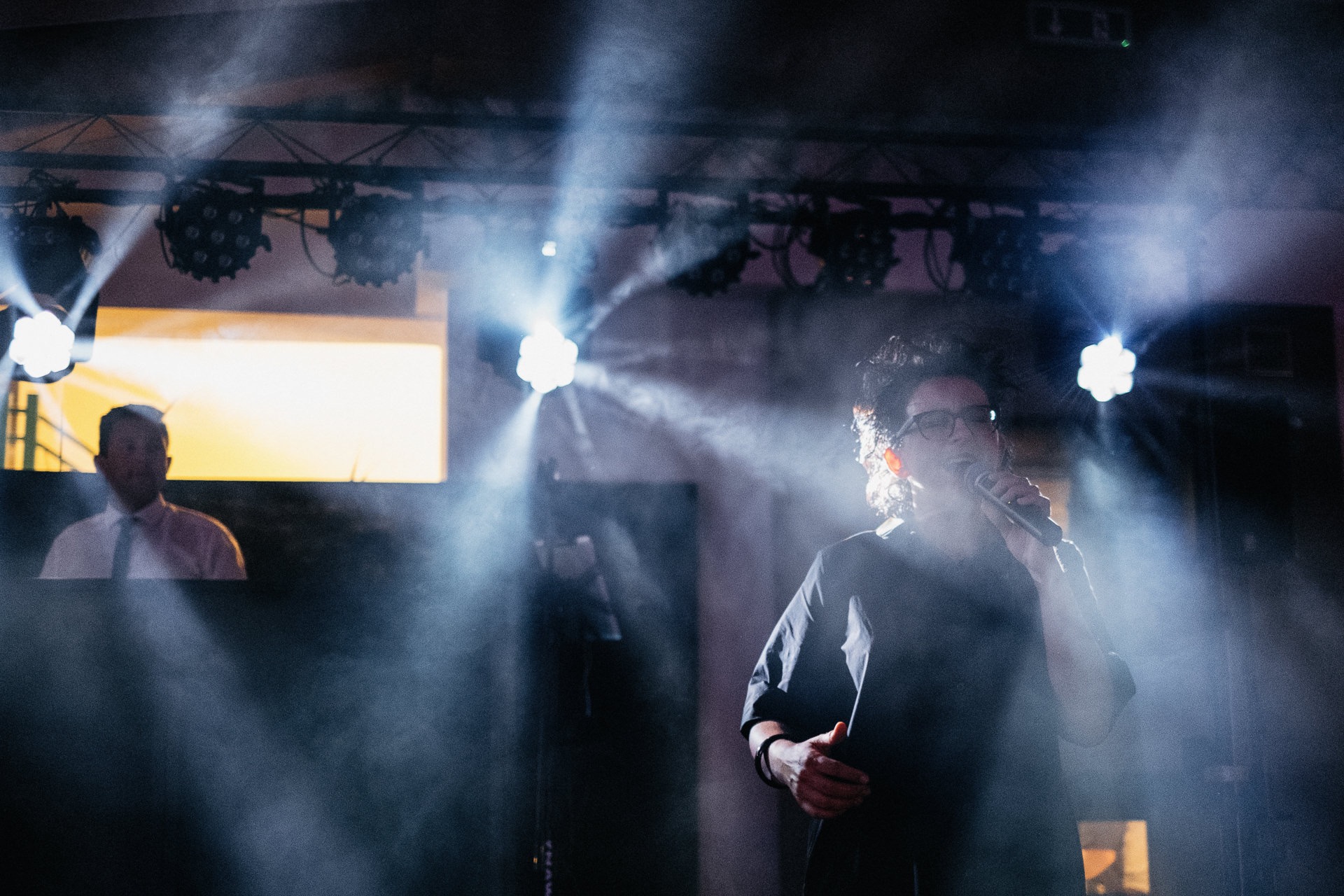 Wokalistka w okularach śpiewa do mikrofonu na scenie oświetlonej reflektorami i wypełnionej dymem, w tle widoczny mężczyzna za ekranem.