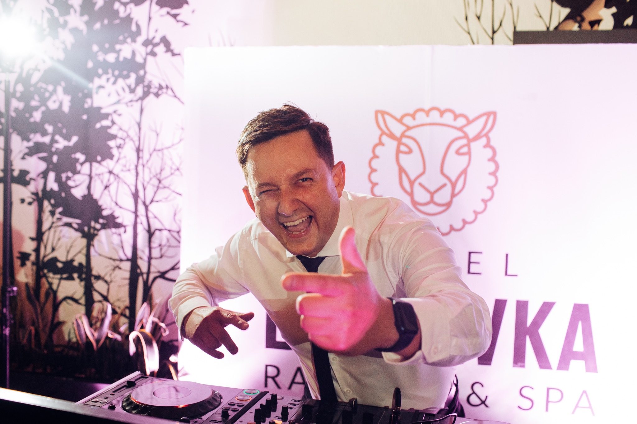 Uśmiechnięty DJ w białej koszuli i krawacie, mruga okiem i wskazuje palcem, grając na sprzęcie DJ-skim na tle ściany z motywem drzew i logiem hotelu.