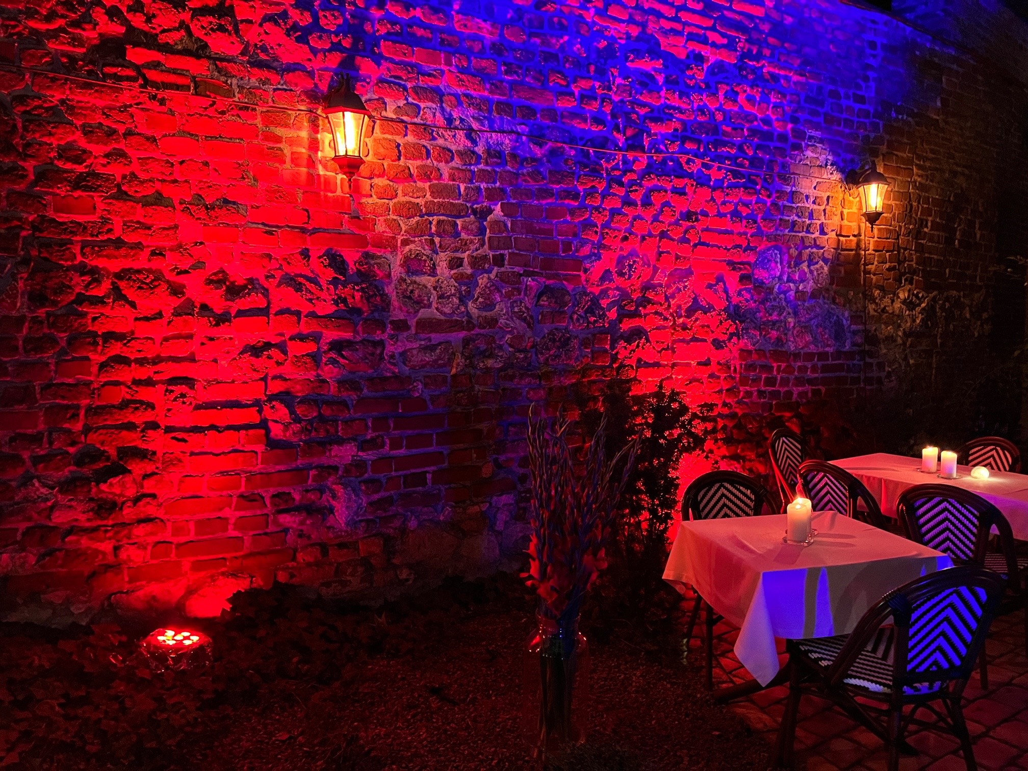 Oświetlona czerwienią ceglana ściana z dwoma kinkietami, stoliki z białymi obrusami i świecami, dekoracyjne oświetlenie podłogowe, oraz bukiet kwiatów w wazonie.