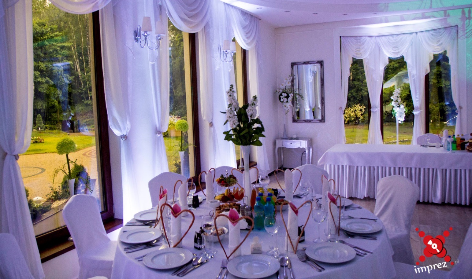 Elegancko udekorowany stół weselny z białymi krzesłami, serwetkami w kształcie serc i kwiatami w wazonie, na tle okien z białymi zasłonami i widokiem na ogród