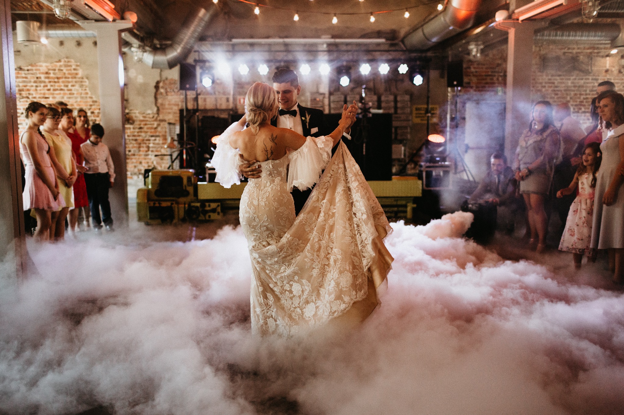 Para młoda tańczy pierwszy taniec w chmurze dymu na tle ceglanej ściany i oświetlenia scenicznego, goście obserwują.