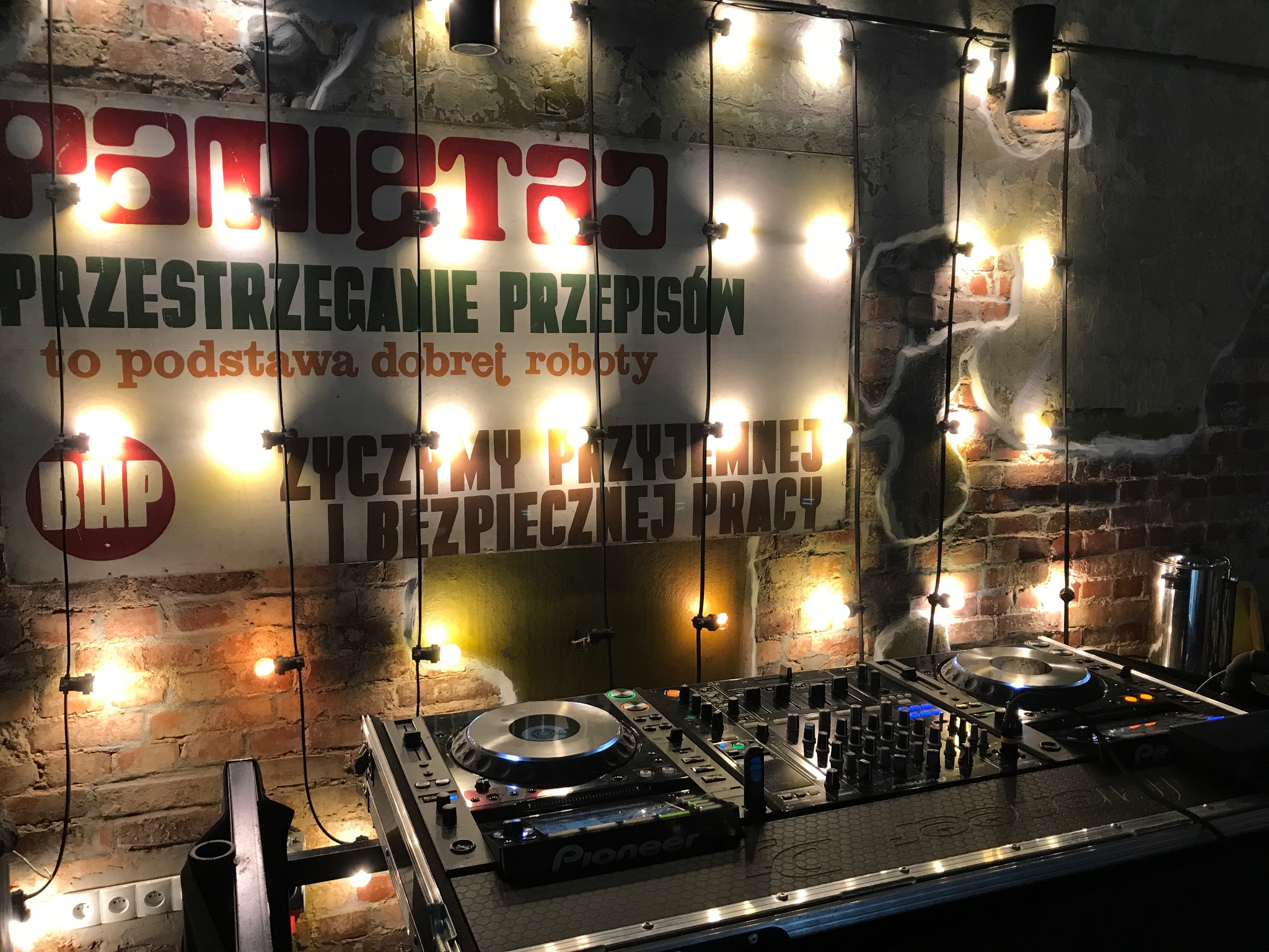 Stanowisko DJ-a z mikserem Pioneer, na tle ceglanej ściany z plakatem BHP i dekoracyjnymi żarówkami.