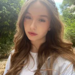 Portret młodej kobiety z długimi, falowanymi brązowymi włosami na tle zieleni, z delikatnym makijażem i błyszczącymi akcentami na twarzy.