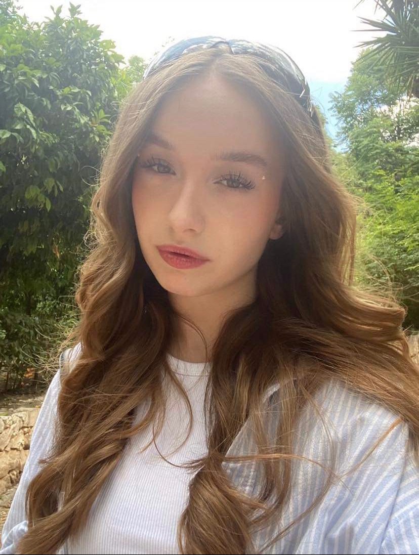 Portret młodej kobiety z długimi, falowanymi brązowymi włosami na tle zieleni, z delikatnym makijażem i błyszczącymi akcentami na twarzy.