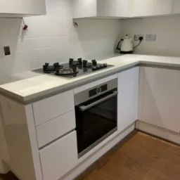 Nowoczesna kuchnia z białymi, lakierowanymi szafkami, marmurowym blatem, płytą gazową i piekarnikiem Bosch, białe płytki na ścianie, drewniana podłoga, widoczne kable wystające ze ściany.