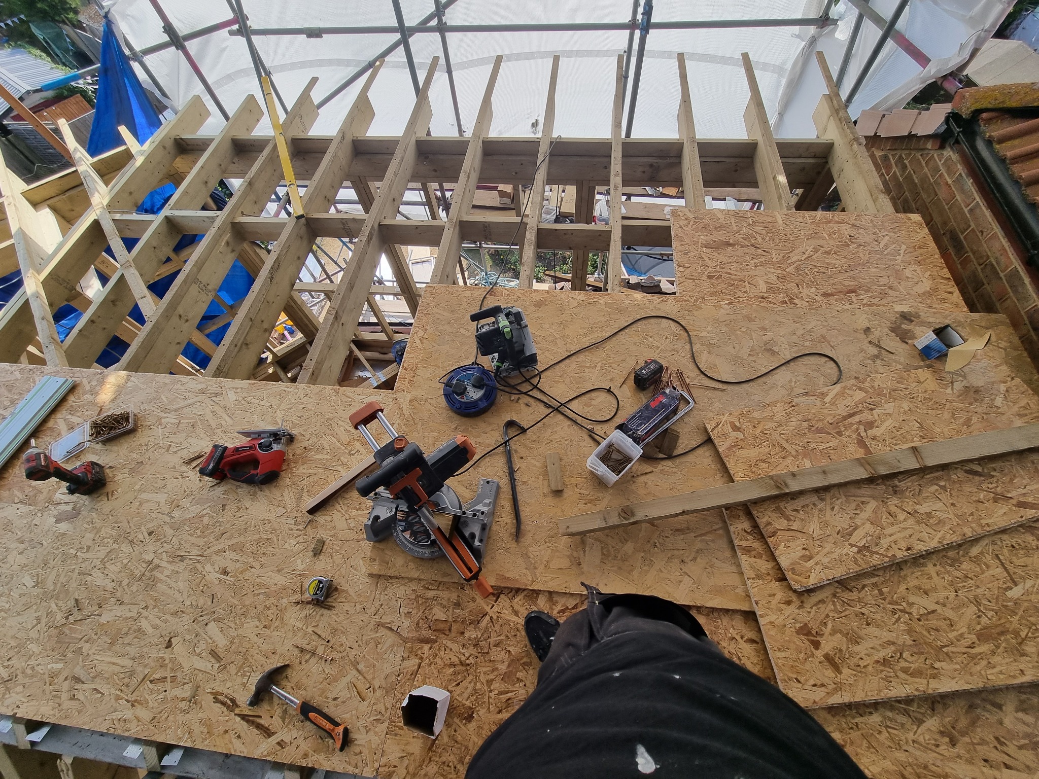 Widok z góry na konstrukcję dachu w trakcie budowy, z widocznymi narzędziami, deskami i płytami OSB, częściowo zakryta rusztowaniem i plandeką, z perspektywy osoby stojącej na dachu.