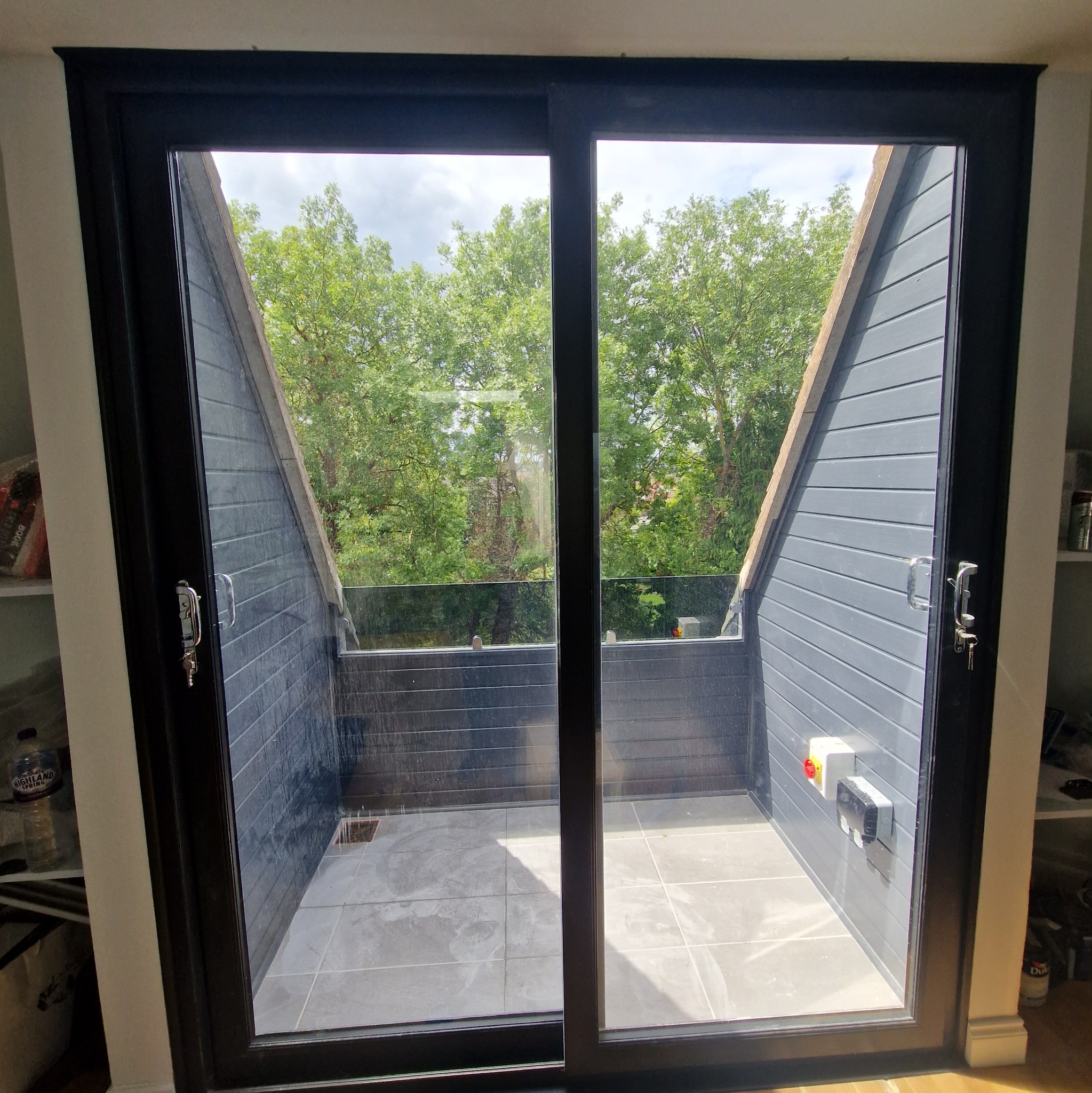 Zadaszony balkon z szarymi panelami ściennymi i płytkami podłogowymi, widok przez przesuwne okno z czarną ramą, zieleń drzew w tle.