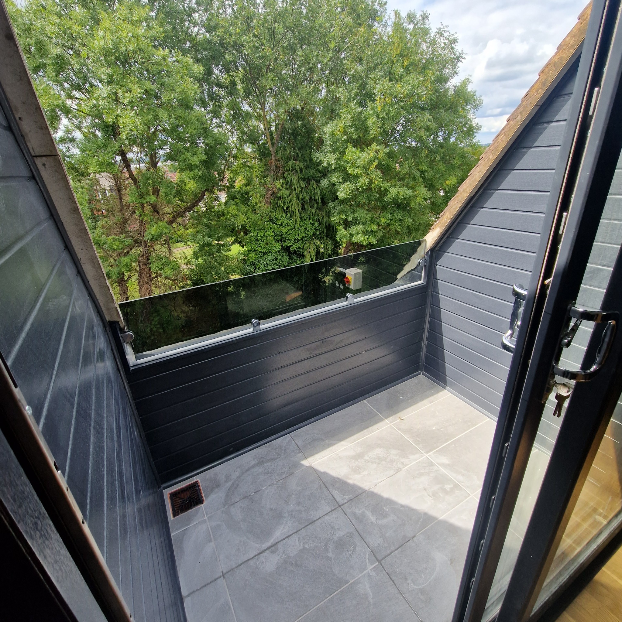 Nowoczesny balkon z szarą okładziną ścian, przeszkloną balustradą i szarą podłogą z płytek, widok na zieleń drzew zza balkonu.