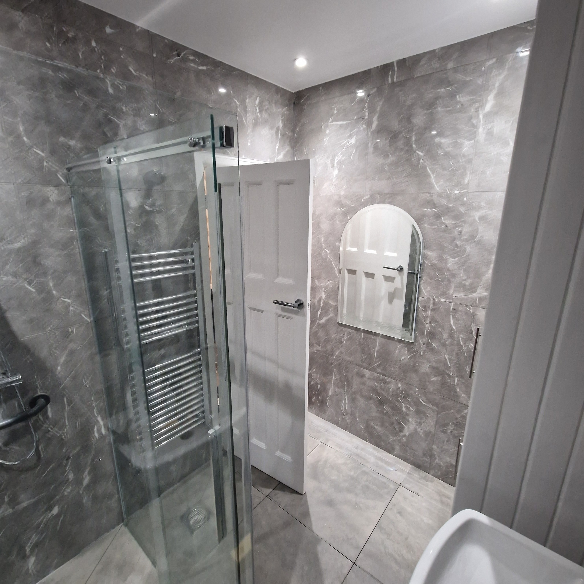 Nowoczesna łazienka z szarymi marmurkowymi płytkami, szklaną kabiną prysznicową z grzejnikiem drabinkowym, białymi drzwiami i owalnym lustrem.