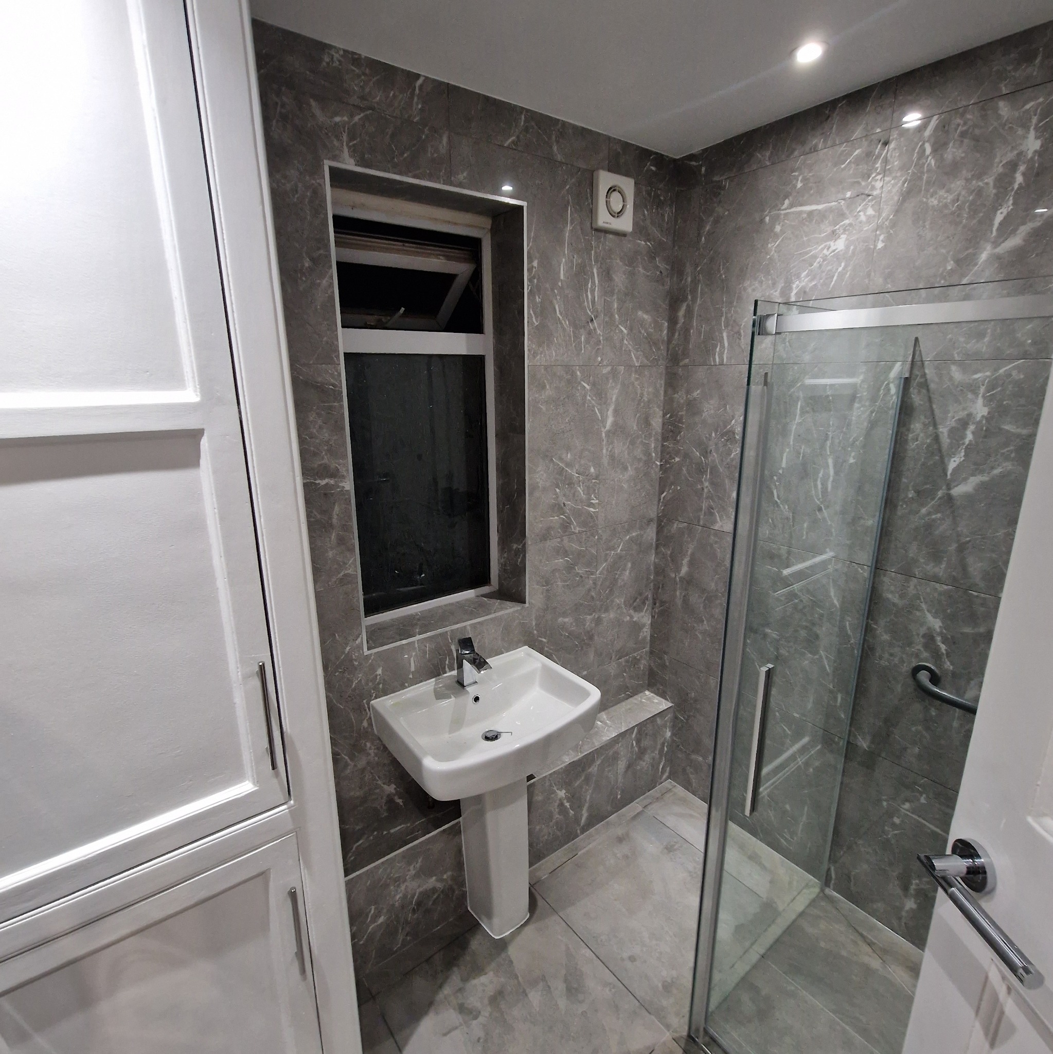 Nowoczesna łazienka z marmurowymi płytkami, białą szafką, umywalką na postumencie i kabiną prysznicową ze szklanymi drzwiami, oświetlona punktowymi lampami sufitowymi.