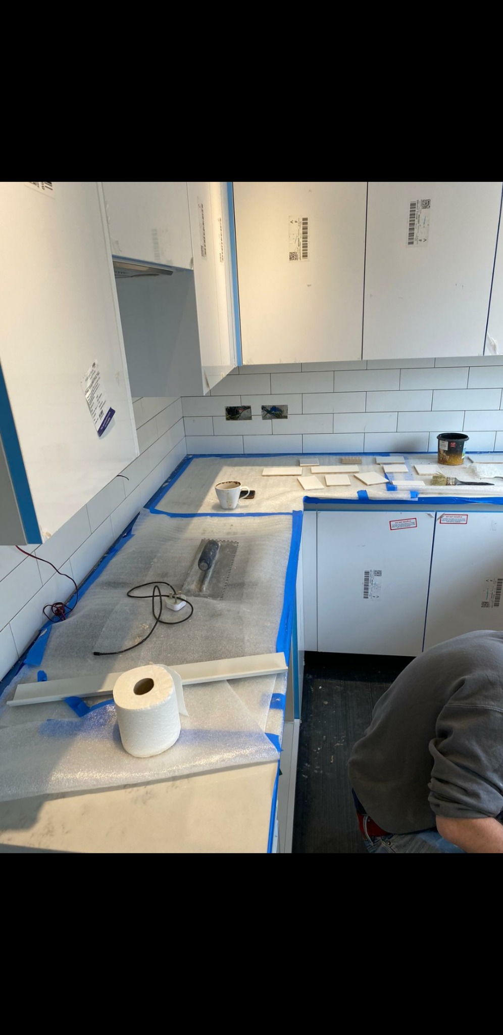 Białe płytki metra ułożone na ścianie nad blatem kuchennym, zabezpieczone niebieską taśmą malarską, z rozłożonymi płytkami i narzędziami do układania oraz kubkiem z kawą.