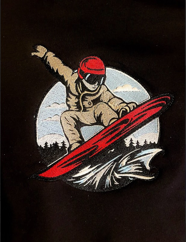 Czarna tkanina z naszywką przedstawiającą snowboardzistę w kasku i goglach, na tle gór i fal, wykonaną techniką haftu.