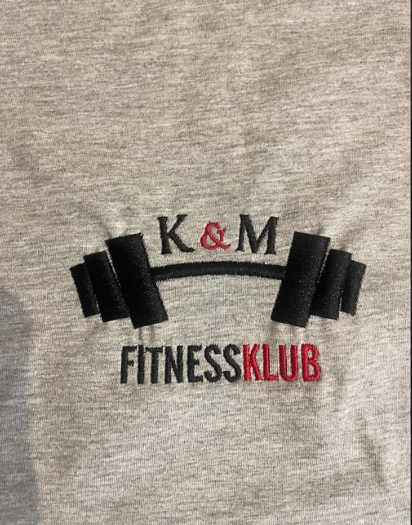 Szara koszulka z wyhaftowanym logo 'K&M FITNESSKLUB' z motywem sztangi, czarne napisy z czerwonymi akcentami.