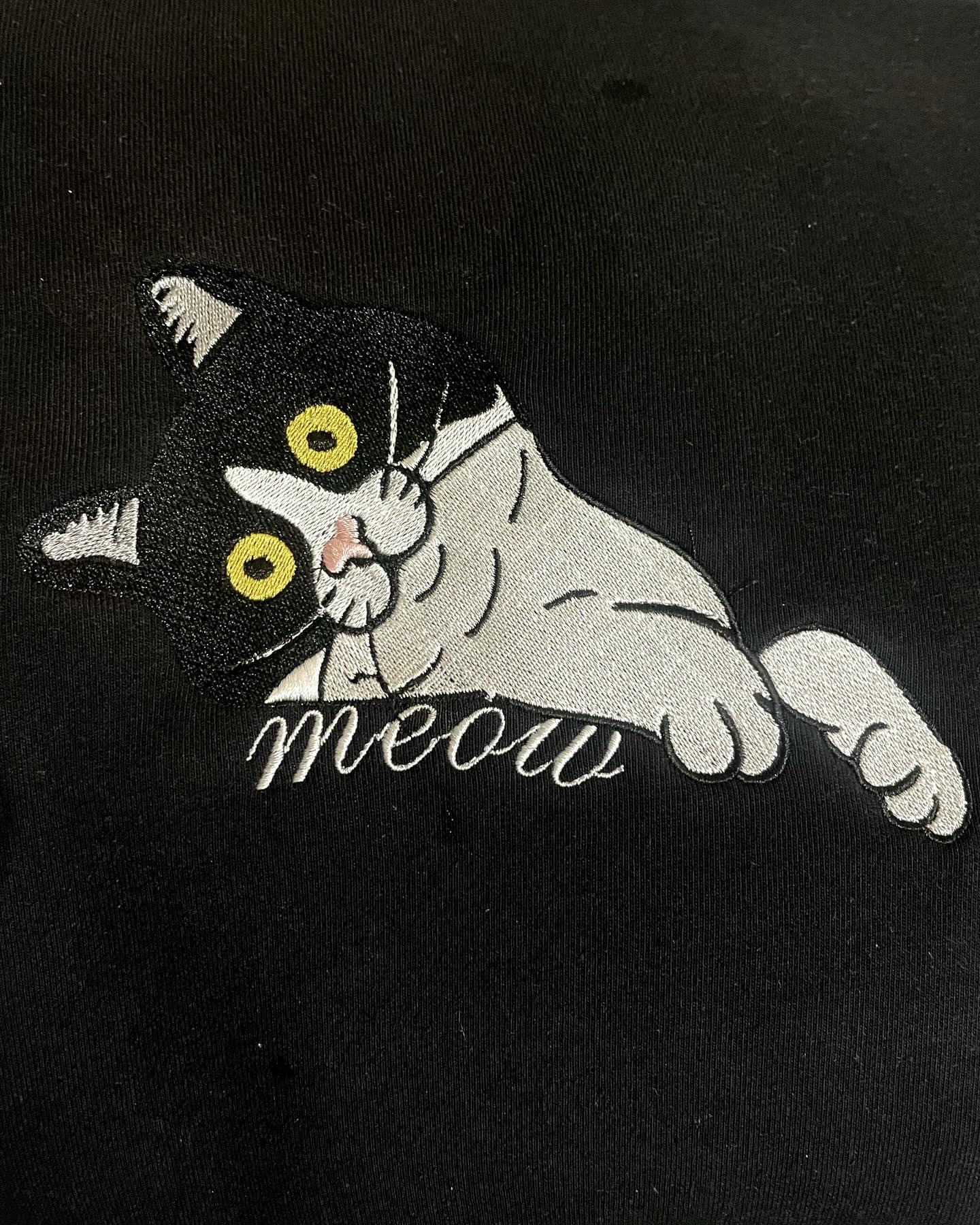 Czarno-biały kot z napisem 'meow' wyhaftowany na czarnej tkaninie, widoczne detale ściegów.