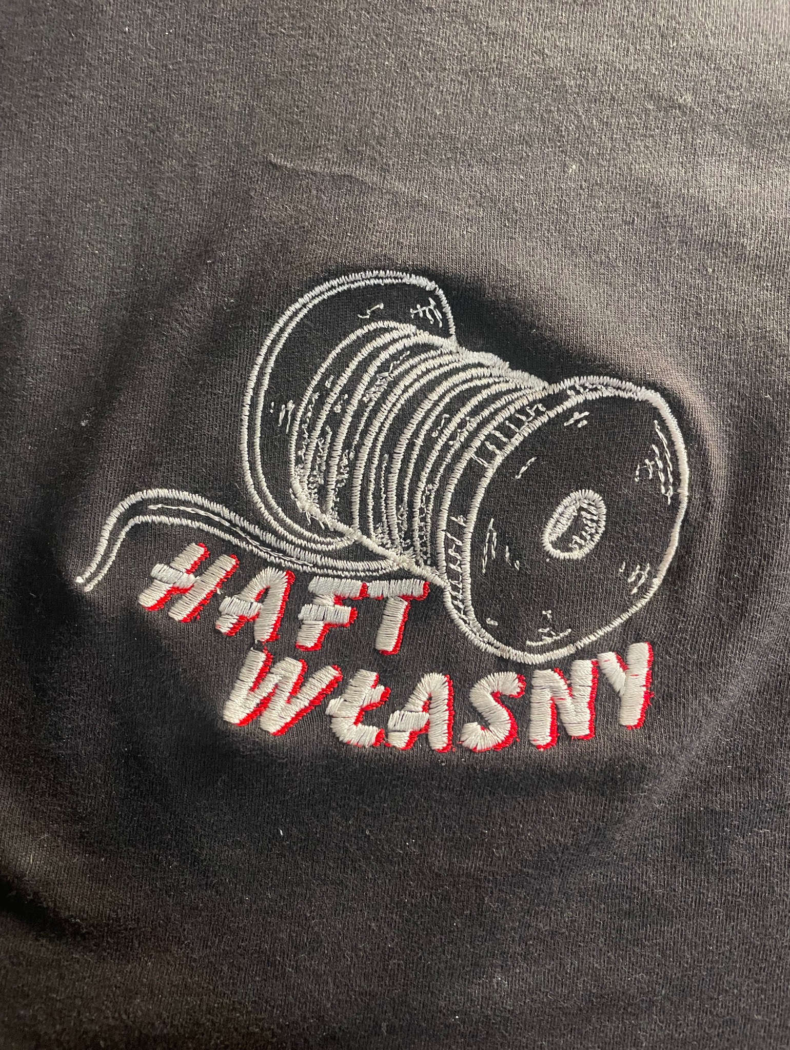 Czarna koszulka z wyhaftowanym motywem szpuli nici w kolorze białym i napisem 'HAFT WŁASNY' w kolorze białym z czerwonym obrysem.