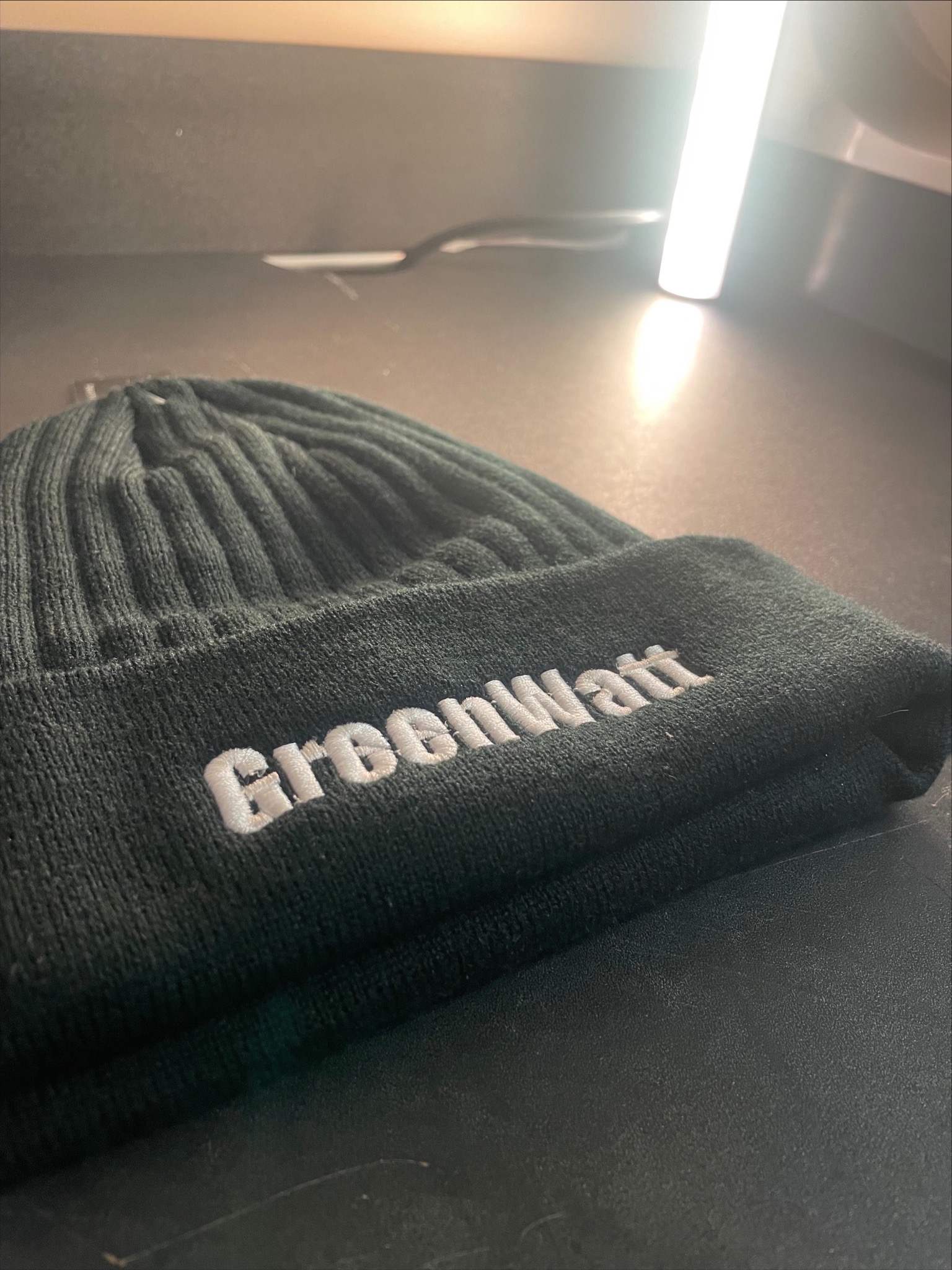 Ciemnozielona, prążkowana czapka zimowa z białym, haftowanym napisem 'Greenwatt' na wywiniętym brzegu, leżąca na ciemnej powierzchni w oświetleniu studyjnym.