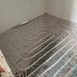 Białe rury instalacji ogrzewania podłogowego ułożone spiralnie na srebrnej folii z czerwoną siatką w rogu pomieszczenia z surową ścianą