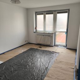 Michał Słodkiewicz - Pokój przygotowany do prac wykończeniowych: podłoga zabezpieczona kartonem i folią, okna i grzejnik osłonięte folią, ściany świeżo pomalowane na biało, widoczne taśmy malarskie.