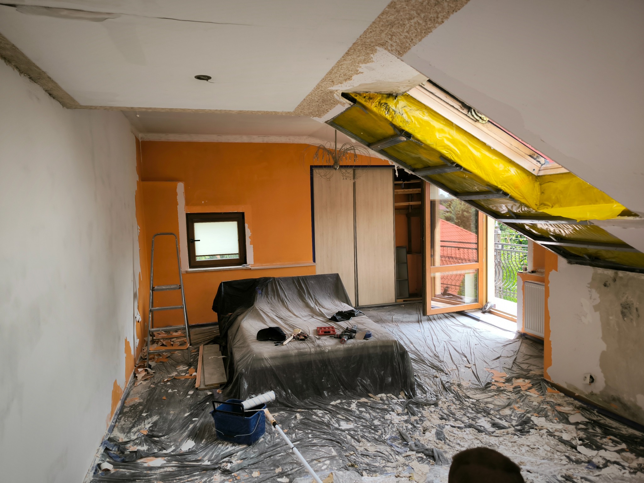 Pokój w trakcie prac wykończeniowych: częściowo pomalowane ściany, zabezpieczone meble folią, widoczne narzędzia i resztki materiałów na podłodze, okno dachowe zabezpieczone żółtą folią.