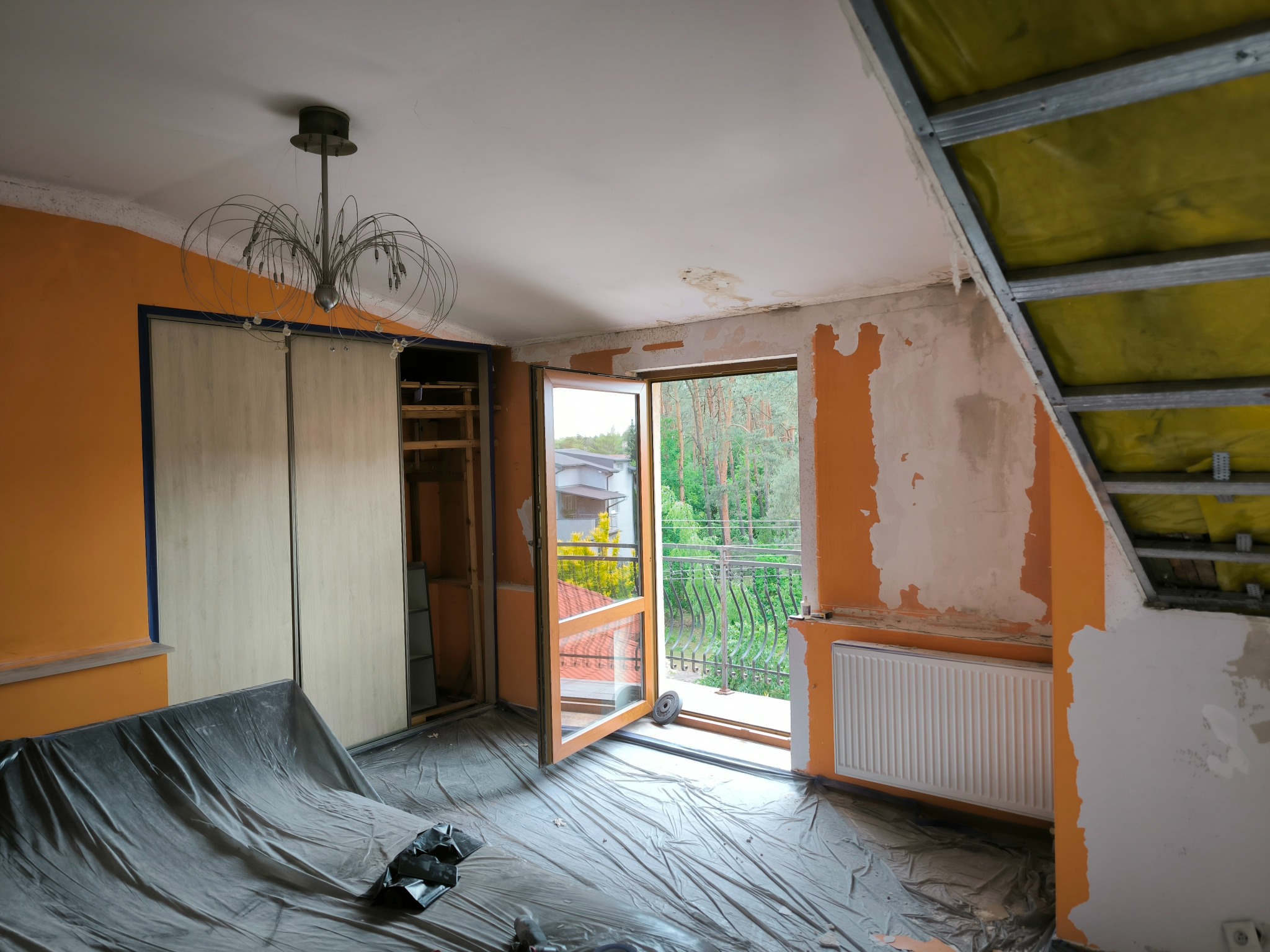 Pokój w trakcie prac przygotowawczych do remontu: zabezpieczona folią podłoga, ściany częściowo odarte z farby, widoczny grzejnik, otwarte drzwi balkonowe z widokiem na zieleń, odsłonięta...