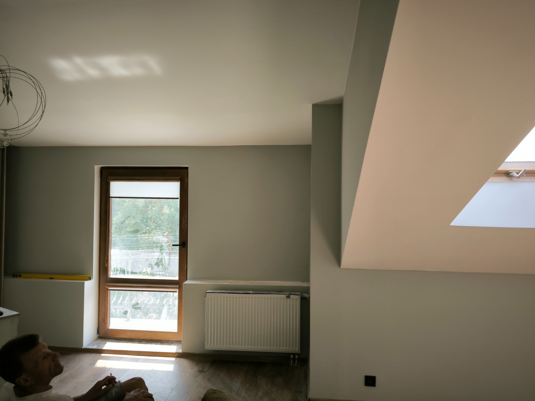 Świeżo pomalowane wnętrze poddasza we Włocławku z oknem dachowym, grzejnikiem i widocznym poziomem na ścianie, w tle odpoczywający mężczyzna.