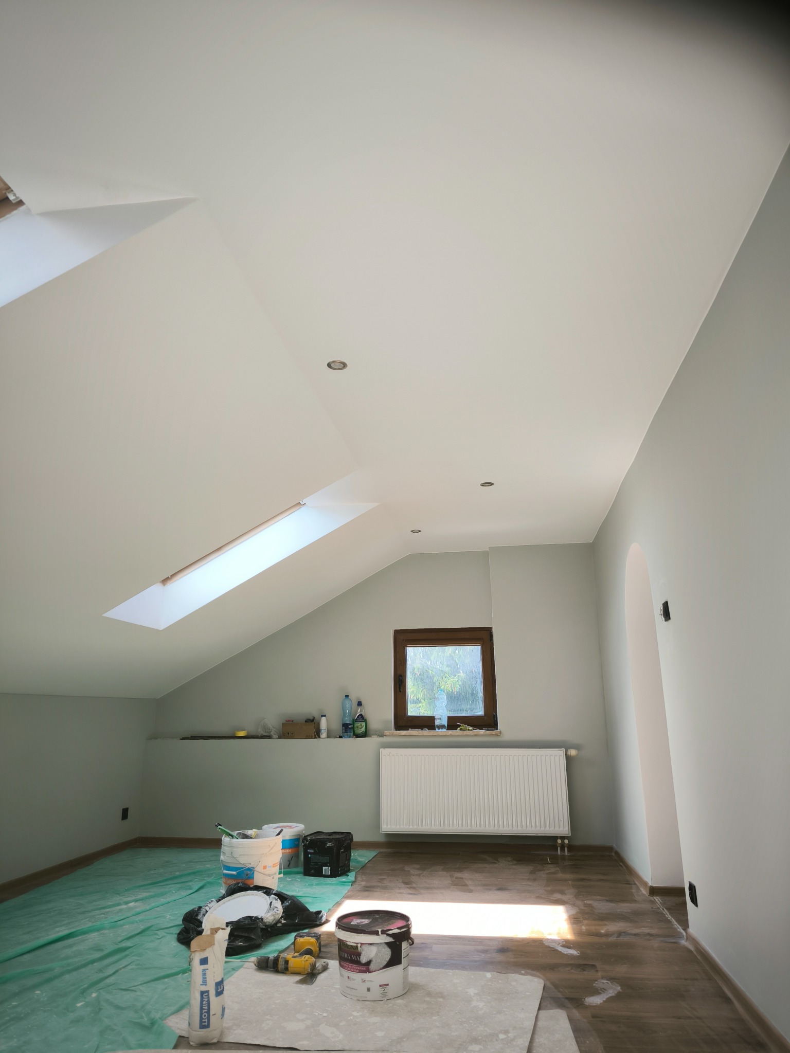 Poddasze po odświeżeniu: Świeżo pomalowane ściany w odcieniach szarości, biały sufit z oknem dachowym i wbudowanymi lampami, zabezpieczona podłoga, widoczne wiadra z farbą, szpachla Knauf Uniflott...