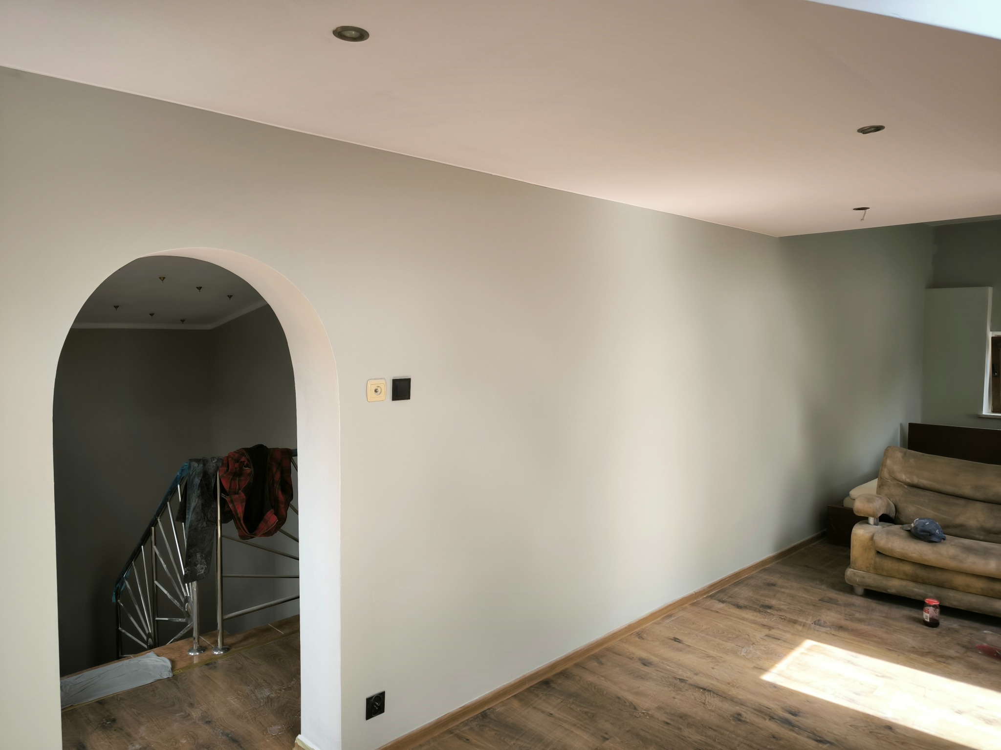 Wnętrze po odświeżeniu: Ściany pomalowane na jasny odcień szarości, drewniana podłoga, widoczny łuk prowadzący do schodów z metalową balustradą, gniazdka elektryczne na ścianie, częściowo zakryty...