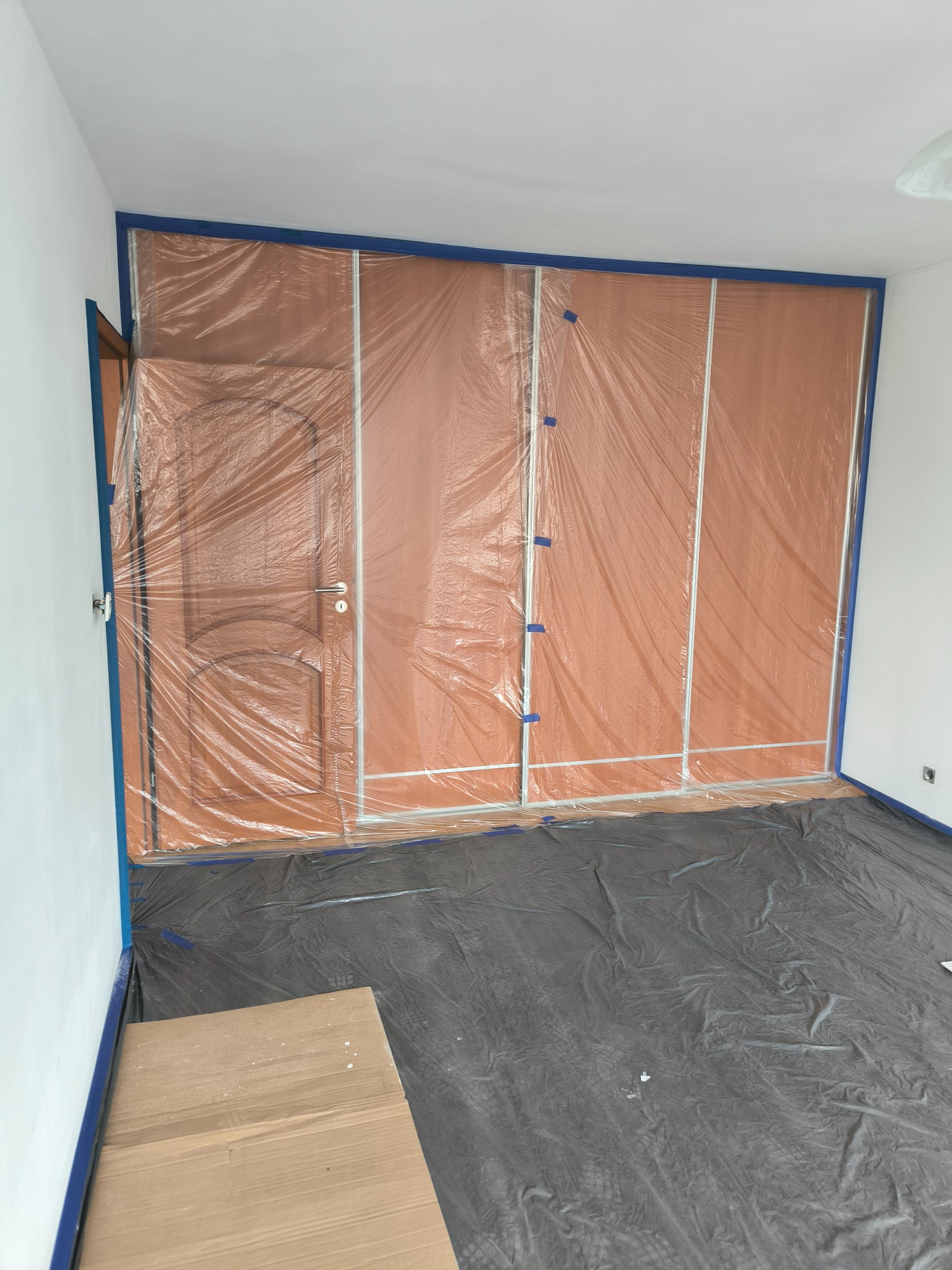 Pokój zabezpieczony folią malarską i taśmą przed malowaniem ścian, widoczna zakryta podłoga i drzwi.