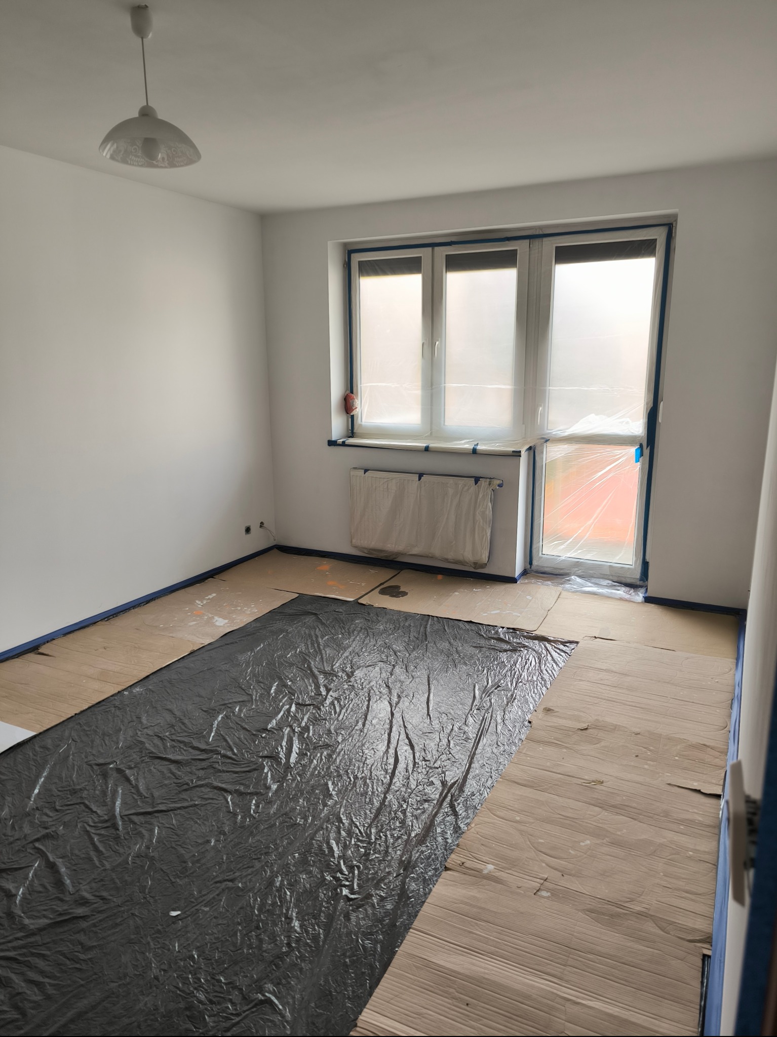 Pokój przygotowany do prac wykończeniowych: podłoga zabezpieczona kartonem i folią, okna i grzejnik osłonięte folią, ściany świeżo pomalowane na biało, widoczne taśmy malarskie.