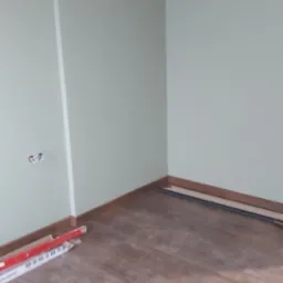 Pok&oacute;j po remoncie: świeżo pomalowane ściany w odcieniach zieleni, ułożone panele podłogowe imitujące drewno, listwy przypodłogowe zamontowane, widoczne puszki elektryczne, paczka paneli pod ścianą.