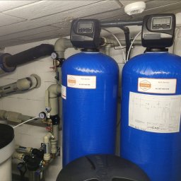 M.R.Service Michał Rowicki - Dwa niebieskie zbiorniki do uzdatniania wody z czarnymi sterownikami, widoczne rury, manometry i elementy instalacji w pomieszczeniu technicznym.