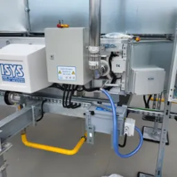 Szczegółowe ujęcie instalacji przemysłowej z rurami, przewodami i skrzynkami sterowniczymi na metalowej konstrukcji. Widoczna żółta rura gazowa, niebieski przewód i białe skrzynki z etykietami.