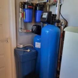 Kompletny system uzdatniania wody z filtrami mechanicznymi, zmiękczaczem i lampą UV, zainstalowany przy ścianie w pomieszczeniu gospodarczym.
