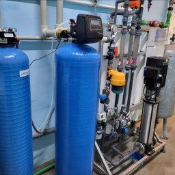 M.R.Service Michał Rowicki - Kompletny system uzdatniania wody z dwoma niebieskimi zbiornikami ciśnieniowymi, pompą Grundfos i licznymi rurami oraz zaworami w pomieszczeniu technicznym.
