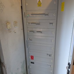 M.R.Service Michał Rowicki - Otwarta, szara skrzynka elektryczna Eaton z bezpiecznikami i oznaczeniami ostrzegawczymi, zamontowana na tle ściany z widocznymi śladami wilgoci.