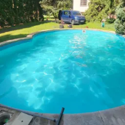 Błękitny basen ogrodowy z czystą wodą w słoneczny dzień. Na skraju leży sprzęt do czyszczenia, a w tle widać dom i zaparkowany samochód.