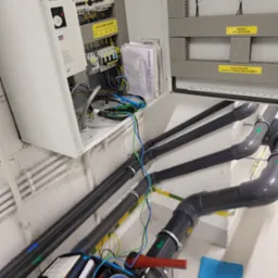 Otwarta szafa elektryczna z rozłącznikiem głównym, widoczne przewody, bezpieczniki, dokumentacja techniczna, miernik elektryczny w niebieskiej torbie i rury kanalizacyjne w tle.