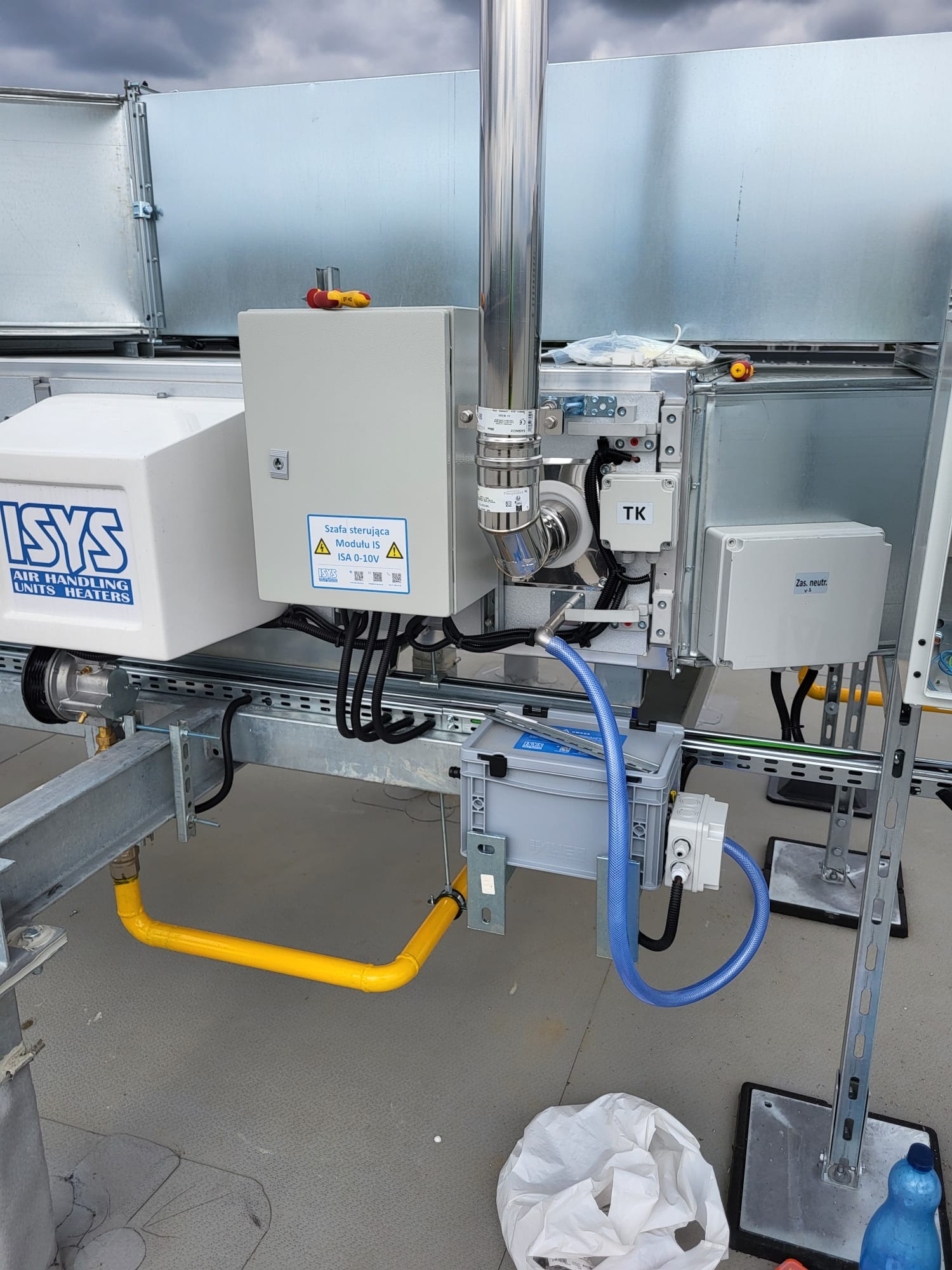 Szczegółowe ujęcie instalacji przemysłowej z rurami, przewodami i skrzynkami sterowniczymi na metalowej konstrukcji. Widoczna żółta rura gazowa, niebieski przewód i białe skrzynki z etykietami.