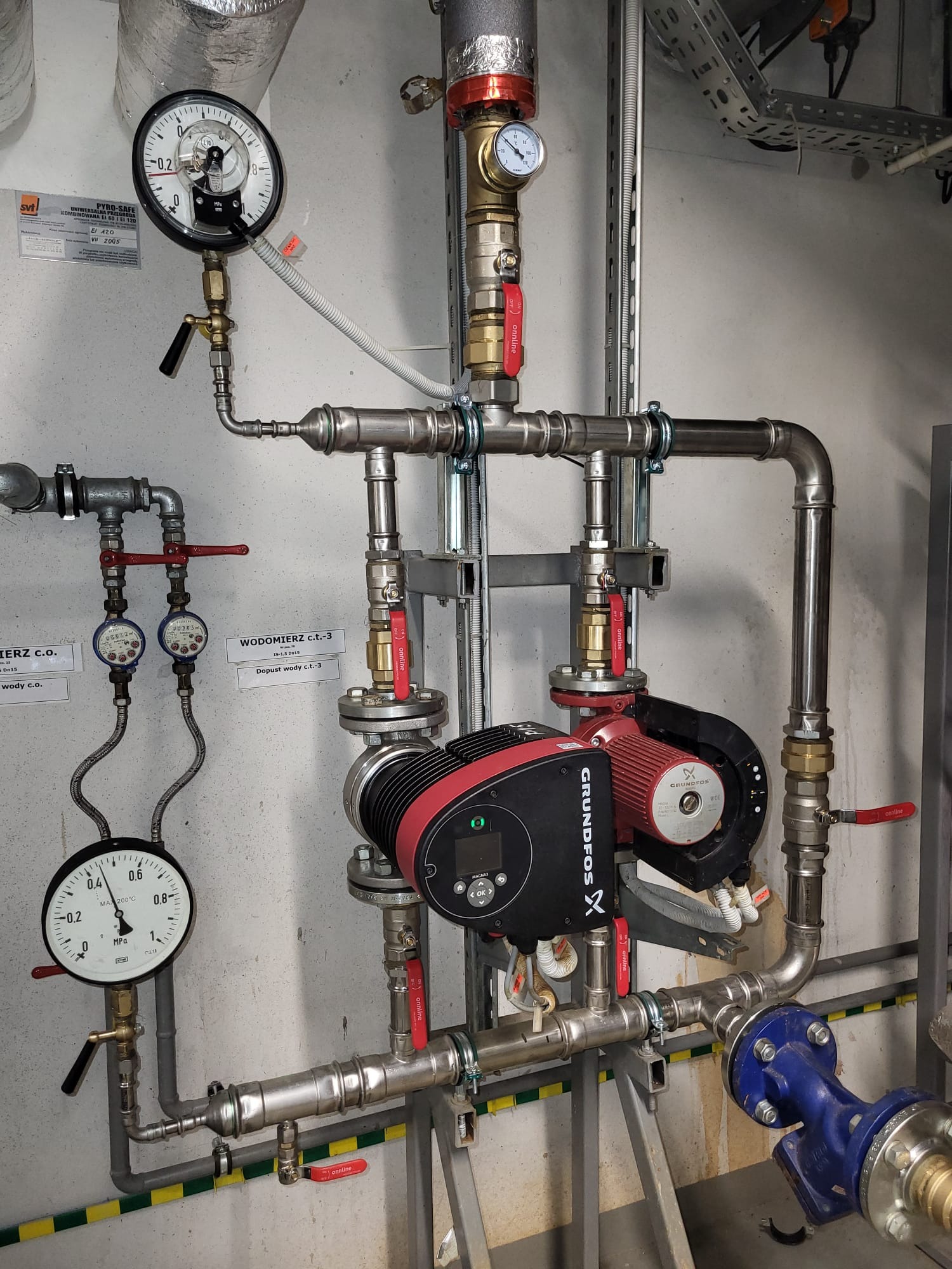 Węzeł cieplny z dwoma pompami obiegowymi Grundfos, manometrami, orurowaniem ze stali nierdzewnej i zaworami kulowymi z czerwonymi rączkami, widoczne wodomierze i tabliczki informacyjne na ścianie.