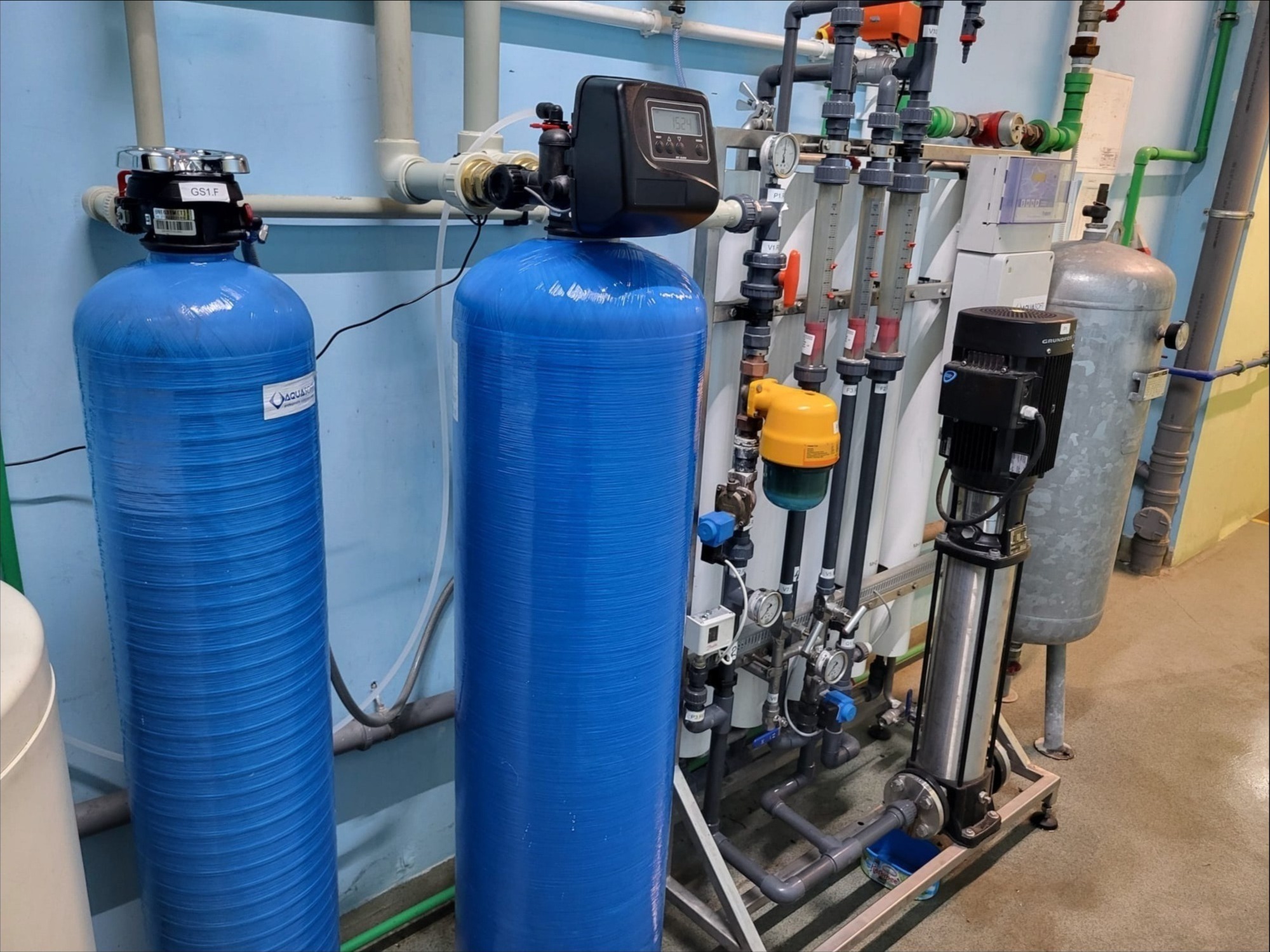 Kompletny system uzdatniania wody z dwoma niebieskimi zbiornikami ciśnieniowymi, pompą Grundfos i licznymi rurami oraz zaworami w pomieszczeniu technicznym.