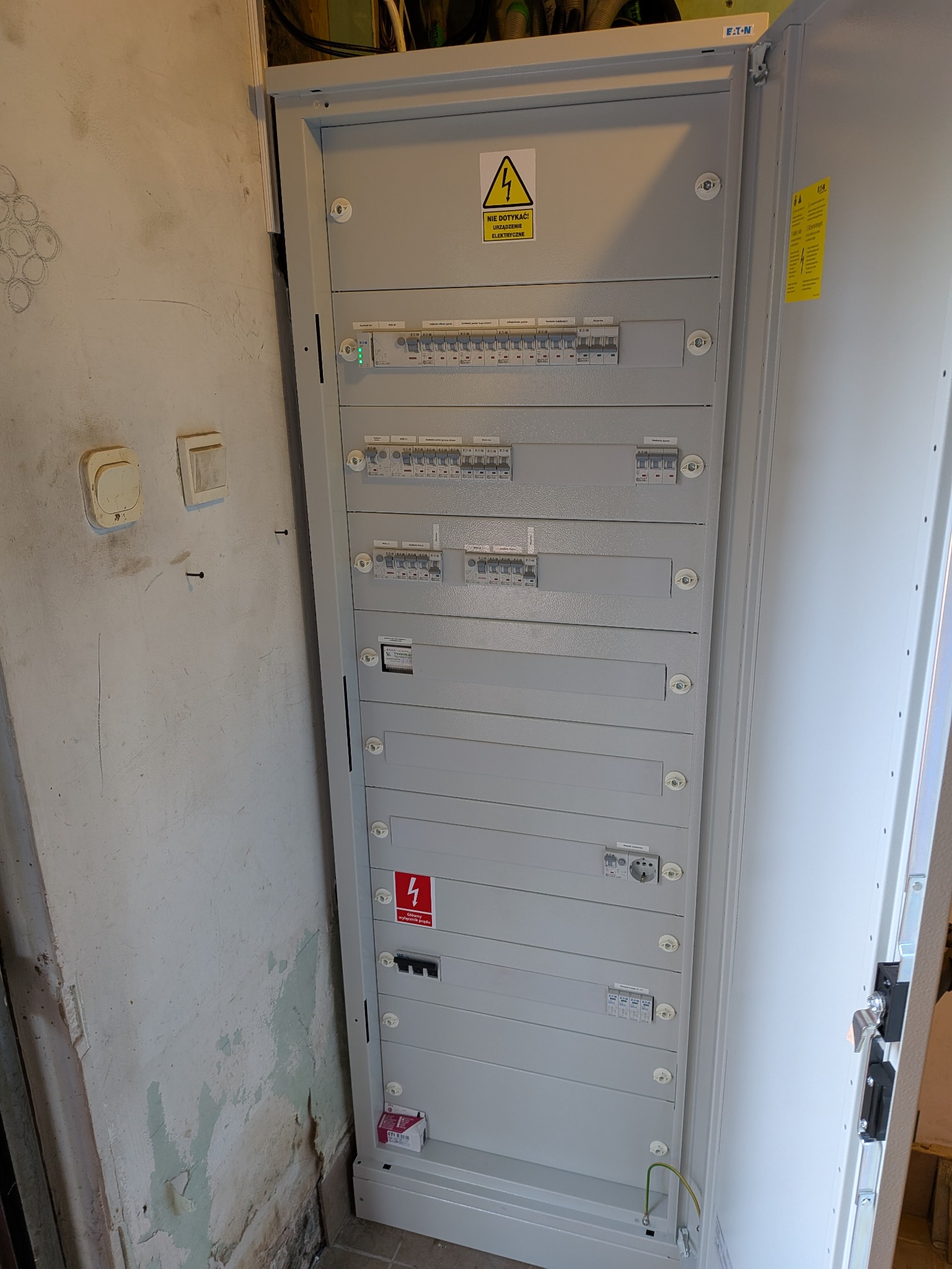 Otwarta, szara skrzynka elektryczna Eaton z bezpiecznikami i oznaczeniami ostrzegawczymi, zamontowana na tle ściany z widocznymi śladami wilgoci.