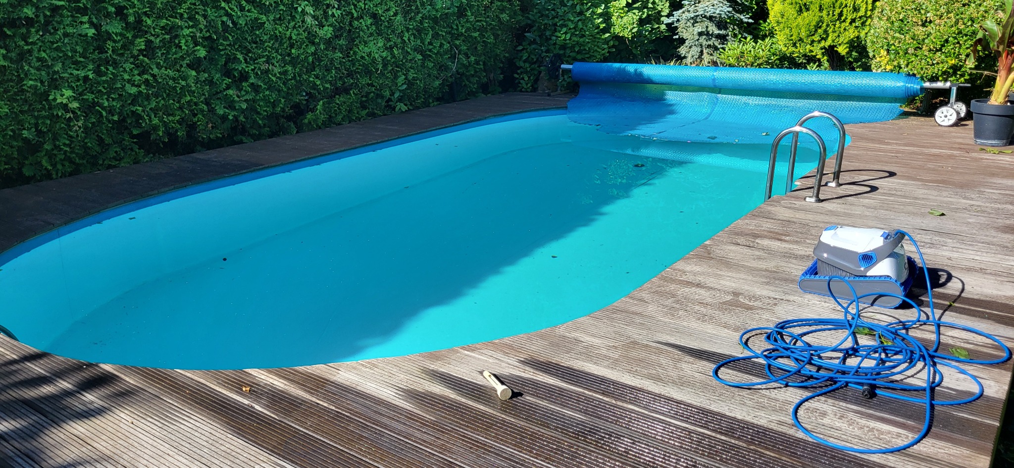 Błękitny basen ogrodowy z drewnianym tarasem, częściowo przykryty folią, z robotem czyszczącym obok drabinki i żywopłotem w tle.