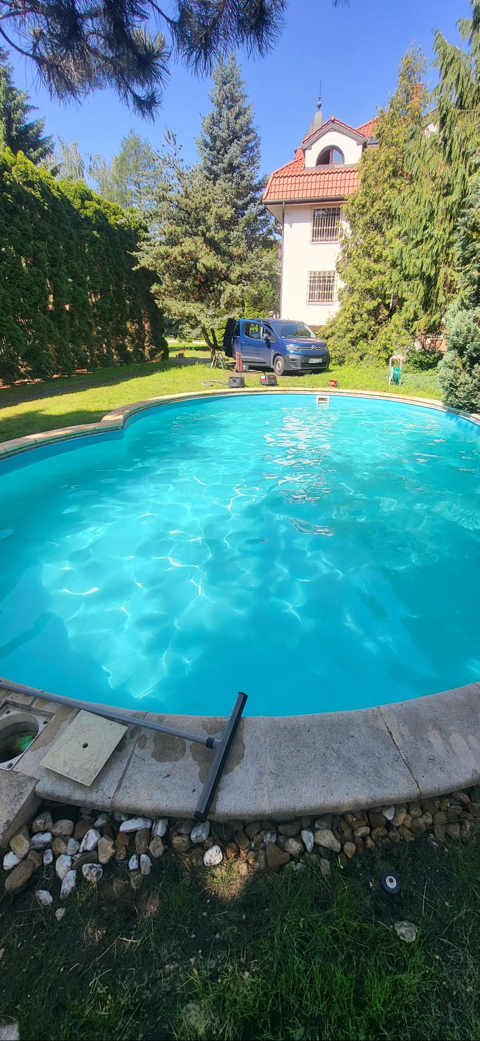 Błękitny basen ogrodowy z czystą wodą w słoneczny dzień. Na skraju leży sprzęt do czyszczenia, a w tle widać dom i zaparkowany samochód.