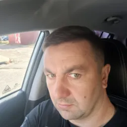 Mężczyzna w czarnej koszulce siedzi w samochodzie, widoczna szyba, fotel i fragment deski rozdzielczej.