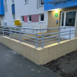 Zewnętrzna balustrada ze stali nierdzewnej, zabezpieczająca podjazd dla osób niepełnosprawnych przy wejściu do budynku mieszkalnego, z numerem 41 nad drzwiami.